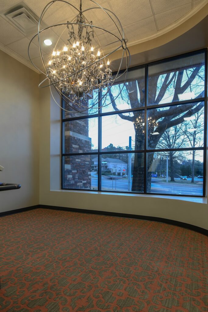 A spacious modern room with a stylish patterned carpet, a large curved window overlooking trees and a street outside, and an ornate chandelier suspended from the ceiling.