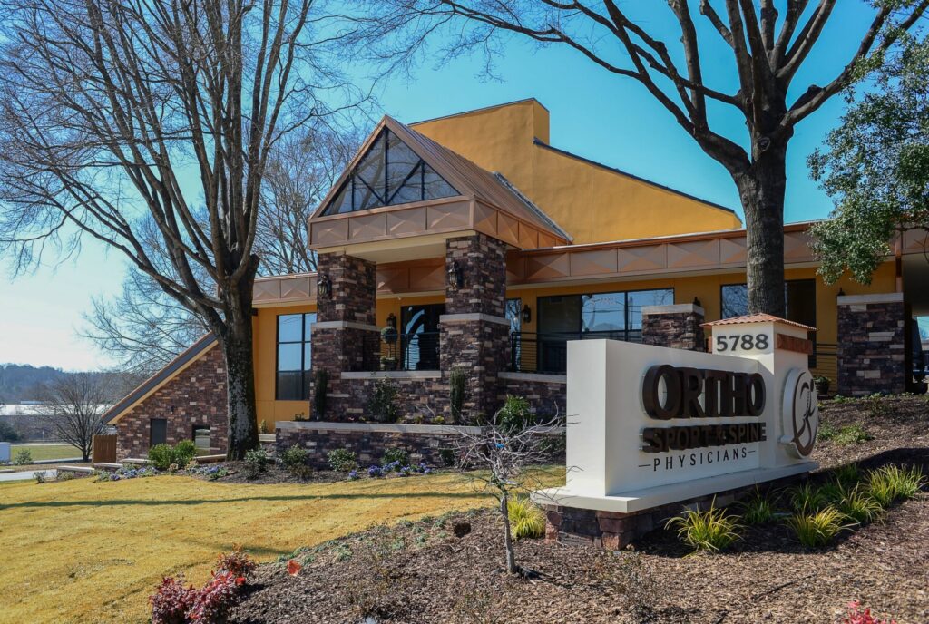 A modern brick and stucco medical office building with a large sign reading Ortho Sport & Spine Physicians and the number 5788, surrounded by landscaped lawns and trees.