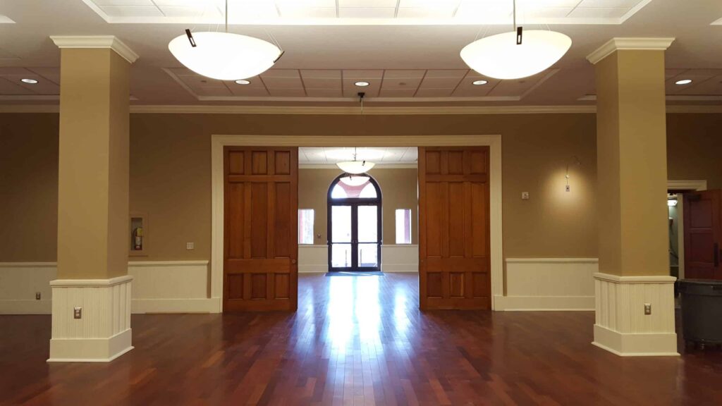 Airy, empty room featuring wooden floors, beige walls, expansive wooden double doors, and three sleek ceiling lights. Sunlight pours through an arched glass door at the far end.