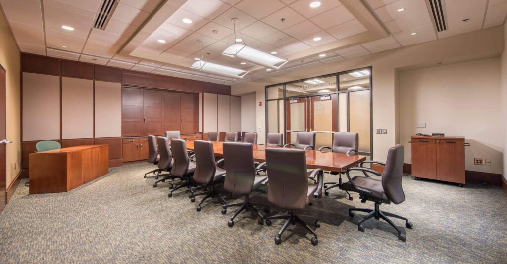 A modern conference room featuring a long wooden table surrounded by twelve gray office chairs, with a carpeted floor and wood accent walls. Overhead lights illuminate the space. A desk sits in the corner, and glass doors are visible at the far end of the room.