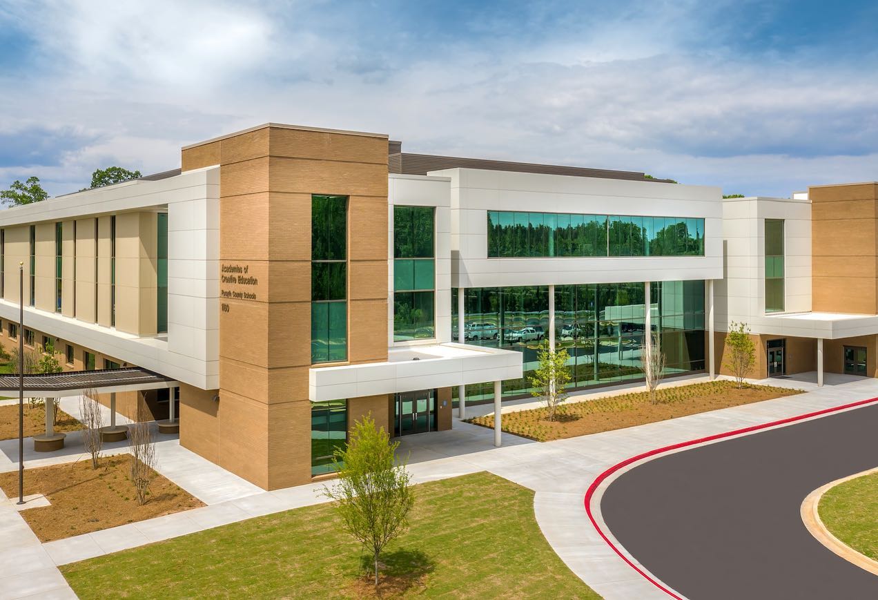 A modern two-story building featuring expansive glass windows and a tan and white exterior, surrounded by green trees and landscaped grounds, with a driveway and parking lot in front.
