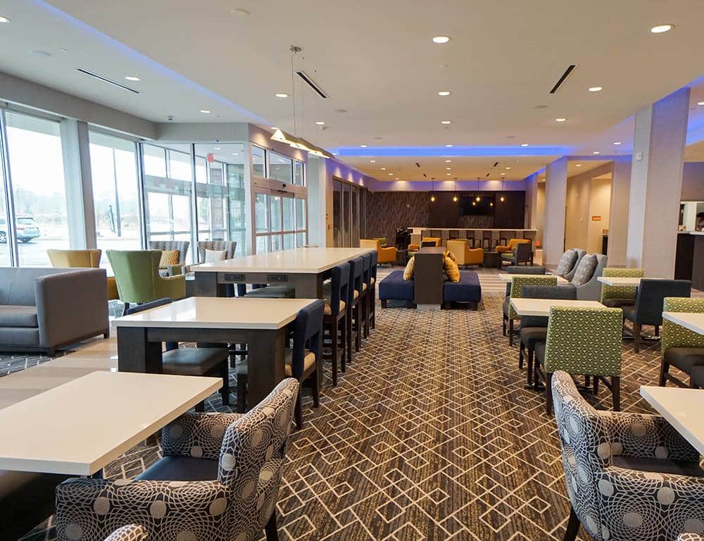 Modern hotel lobby with patterned carpet, various armchairs, sofas, and long tables. Large windows on the left bring in natural light, while ceiling lights and blue accents create a welcoming atmosphere.