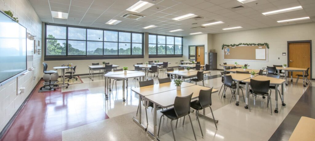 A spacious, modern classroom featuring multiple tables and chairs, large windows filling the room with natural light, a whiteboard at the front, and a bulletin board on the back wall. Each table is decorated with small plants.