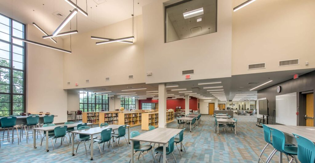 A modern library featuring tall windows, bookshelves, teal chairs, and tables. Geometric lights are suspended from the high ceiling, while a glass-walled upper room looks out over the spacious, well-lit interior.