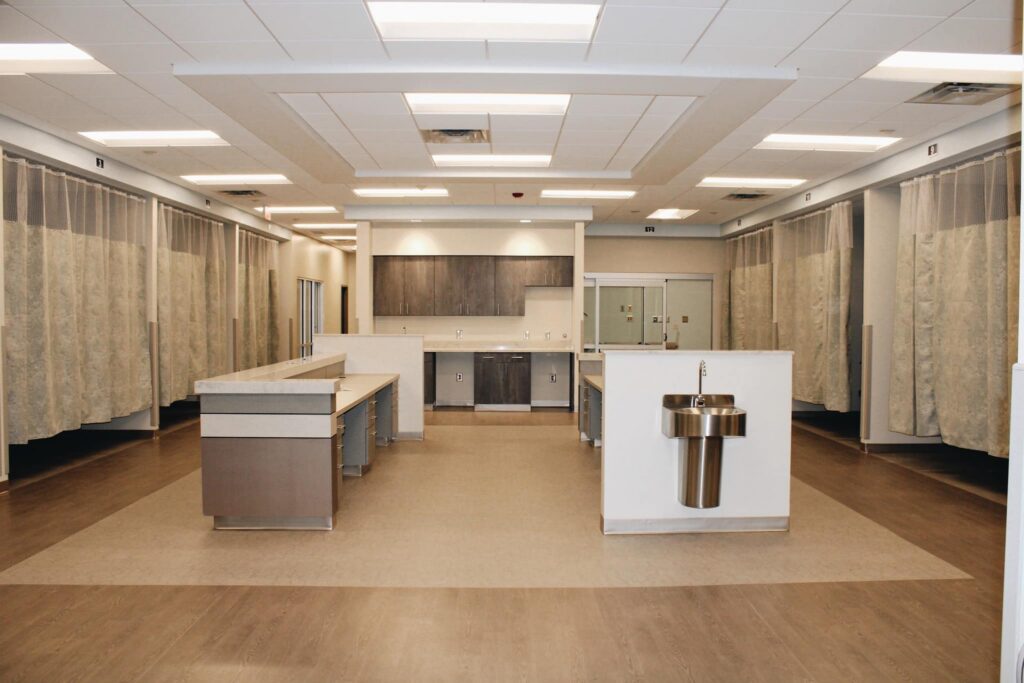 A modern, empty hospital room featuring multiple curtained patient areas, two central stations, a metal sink, and cabinets lining the back wall under bright ceiling lights.