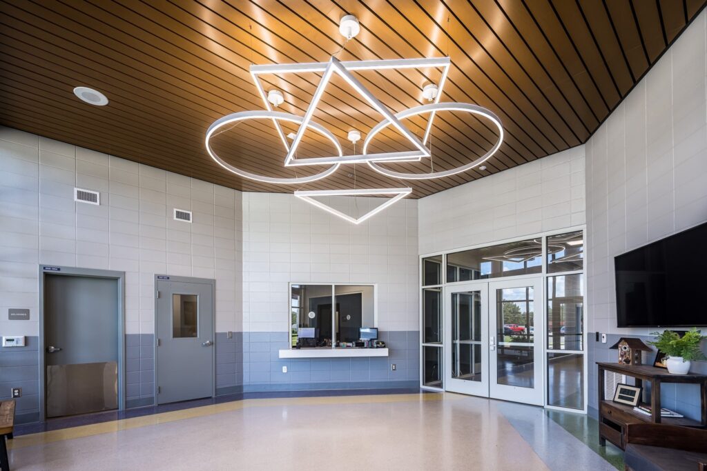 Modern lobby featuring a geometric light fixture, tiled walls, a small reception window, glass entrance doors, and a wood-paneled ceiling. On the right side, there is a TV and a small table with plants.