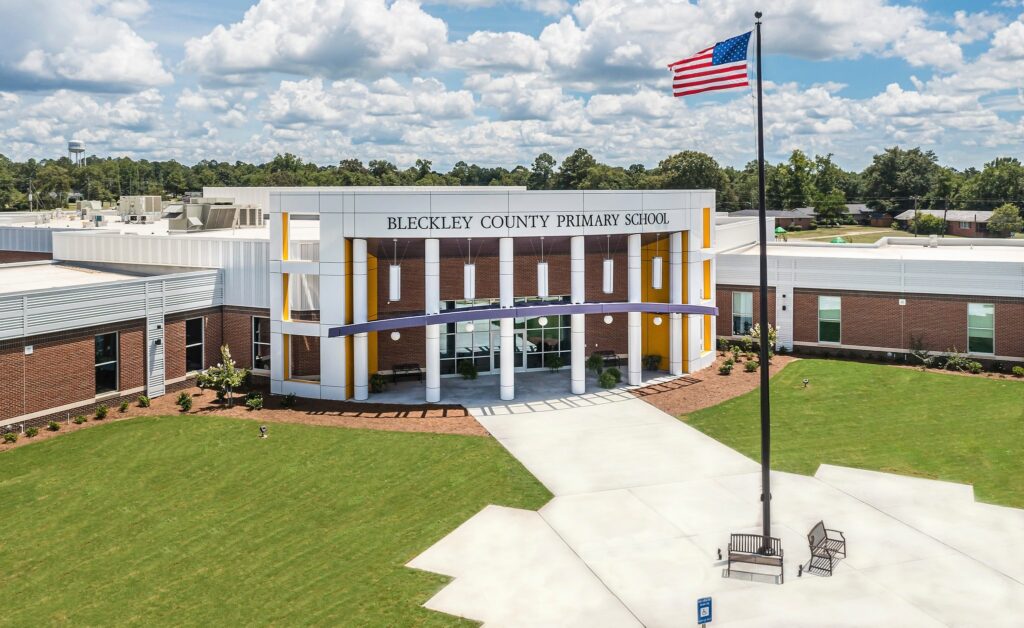 Front entrance of Bleckley County Primary School, featuring a modern building, a U.S. flag on a tall flagpole, and a well-kept green lawn beneath a partly cloudy sky.