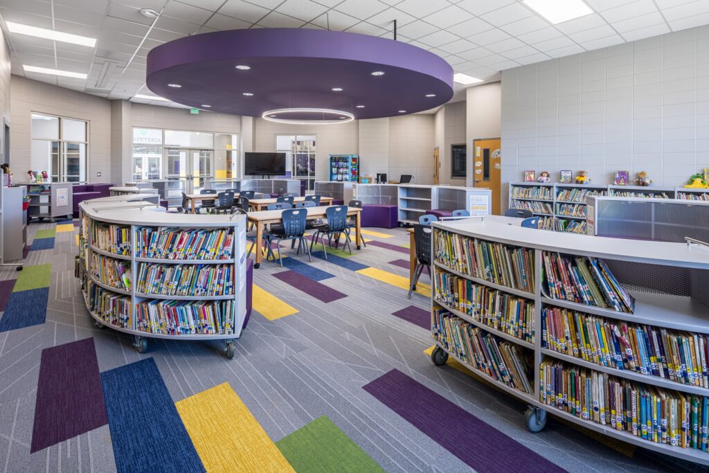 A modern library with curved bookshelves, colorful carpet tiles, tables, and chairs. A circular purple ceiling feature sits above the central area. Shelves are filled with books and decorative items.