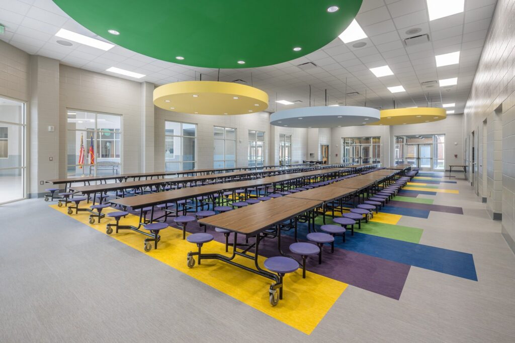 A modern school cafeteria features long tables with round blue seats attached, vibrant geometric patterns on the floor, and oversized circular ceiling lights in shades of green, yellow, and blue. The bright area is filled with natural light from numerous windows.