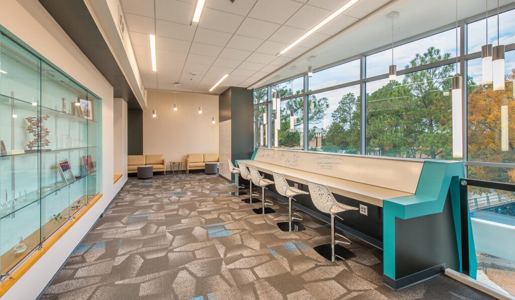 Contemporary study space featuring a patterned carpet, a display case on the left, a tall white counter lined with stools and decorated with math formulas, expansive windows revealing trees outside, and lounge chairs situated in the back corner.