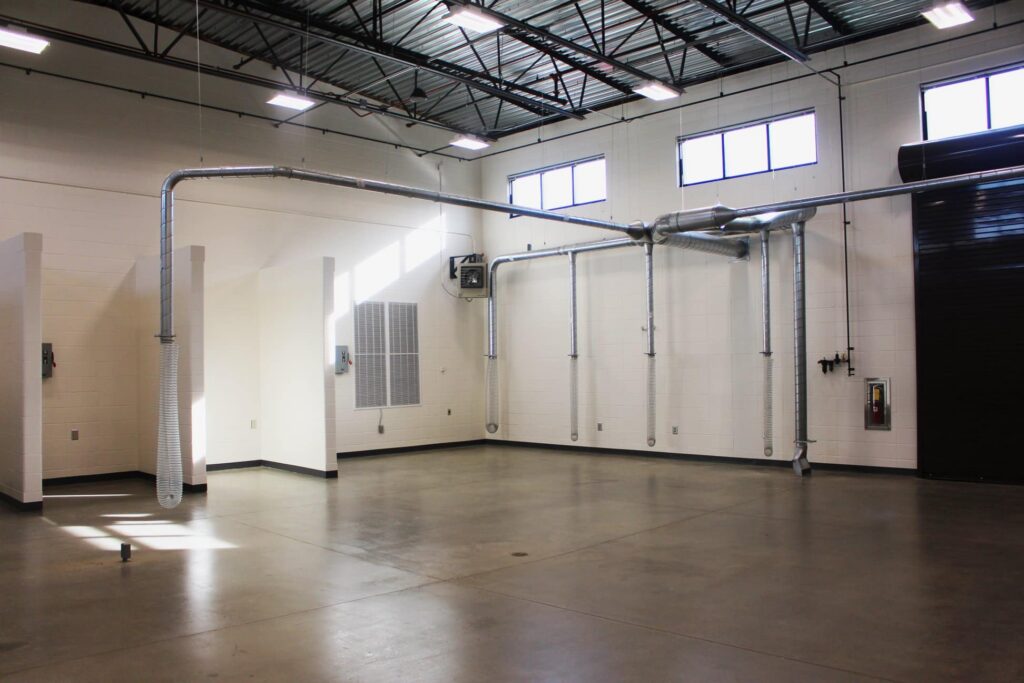 Spacious industrial room featuring polished concrete floors, high ceilings with exposed ductwork, white walls, and several windows that fill the space with natural light.