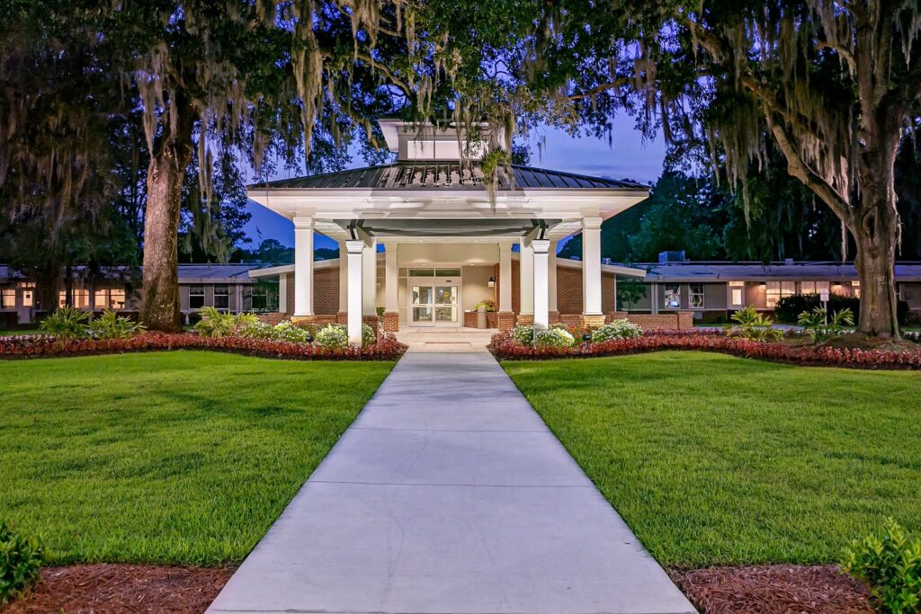 A wide sidewalk stretches toward a brightly illuminated building entrance supported by white columns, bordered by neatly trimmed lawns, colorful flowers, and towering trees draped with hanging moss in the soft light of dusk.