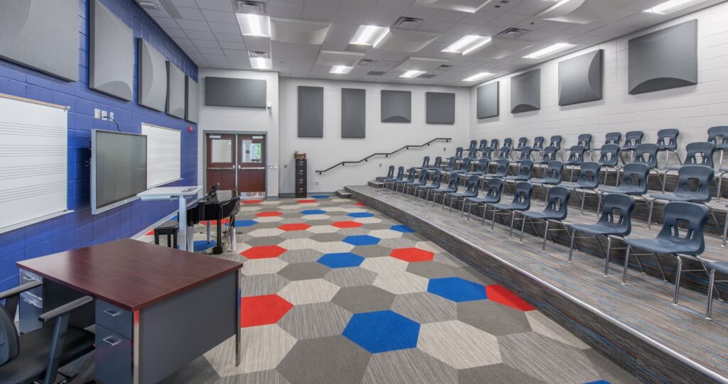 A modern classroom with tiered seating, blue and white walls, a digital whiteboard, a piano, a desk, and a vibrant hexagon-patterned carpet. Acoustic panels are installed on sections of the walls and ceiling.