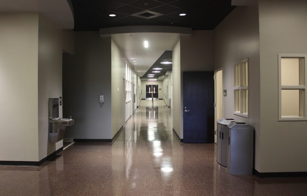A clean, empty hallway in a building with beige walls and shiny brown floors. There is a water fountain, trash bins, and closed doors at the far end. Fluorescent lights run along the ceiling.