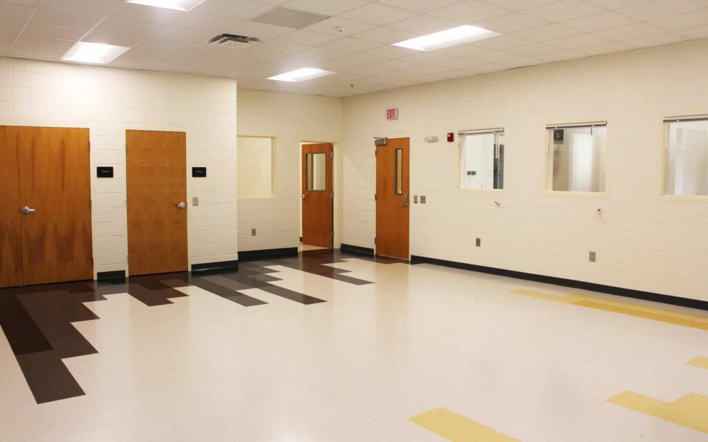 A spacious, uncluttered room featuring white walls, patterned brown and yellow flooring, wooden doors, and multiple windows, suggesting a school or office environment.