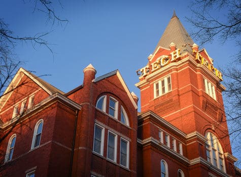 A red brick building with arched windows and a tall clock tower displaying the words TECH TOWER in yellow letters, against a clear blue sky.