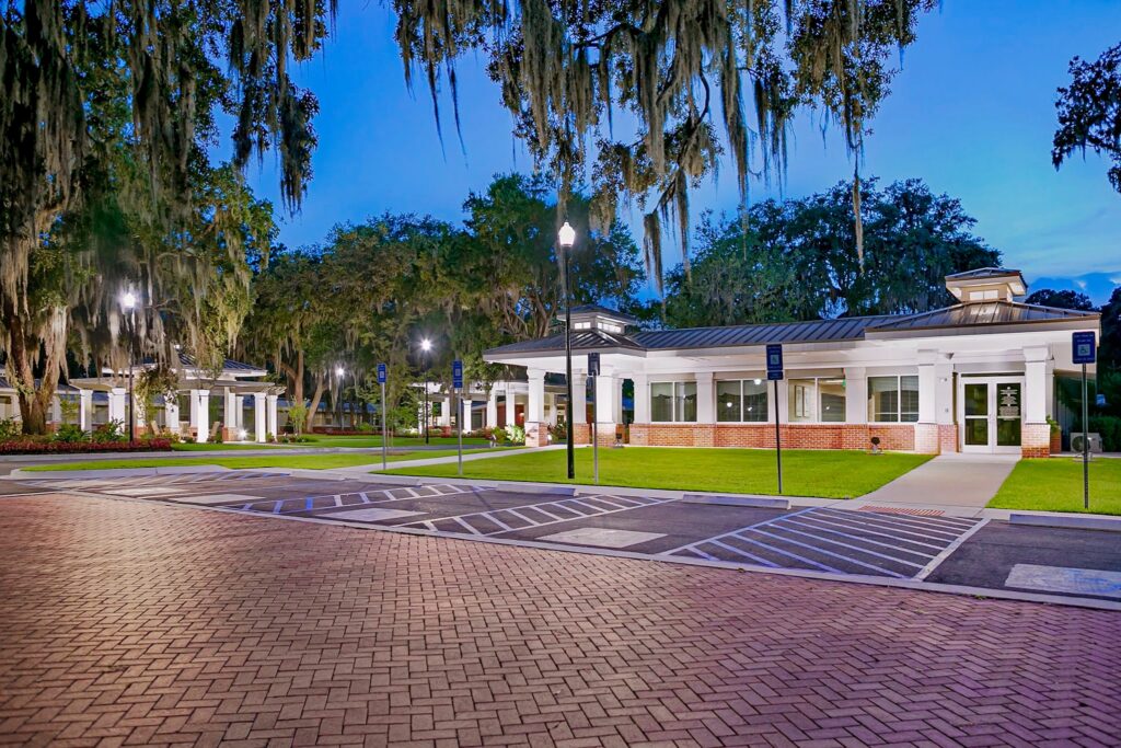 A well-lit building with white columns stands beside an empty brick and asphalt parking lot, surrounded by trees draped in Spanish moss during the evening.