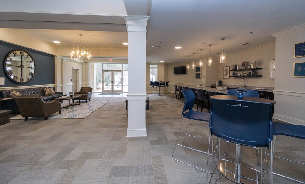 Spacious lounge area featuring modern decor, blue and brown furnishings, a large wall mirror, bar seating, and bright hanging lights. Large windows in the background fill the space with natural light.