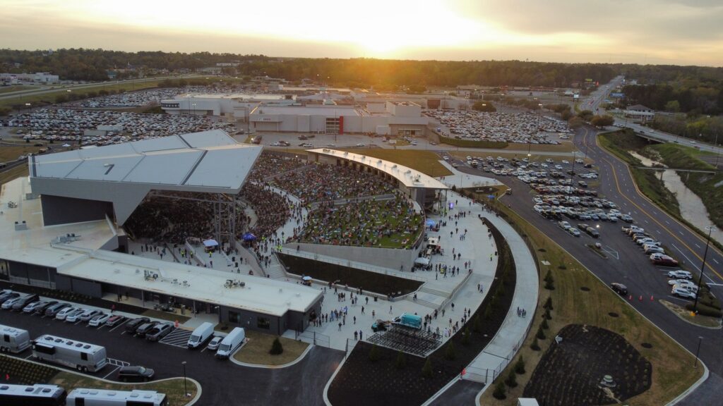 Aerial view of a sprawling outdoor amphitheater filled with people, bordered by parking lots busy with cars, all under the glow of a sunset. The venue features a covered stage and semicircular seating arrangement.