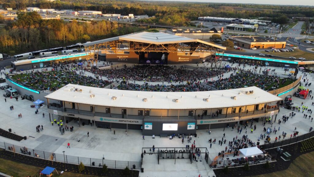 A large outdoor amphitheater filled with people attending a concert, surrounded by parking lots, trees, and nearby buildings under a clear sky. North Gate signage is visible at the entrance.