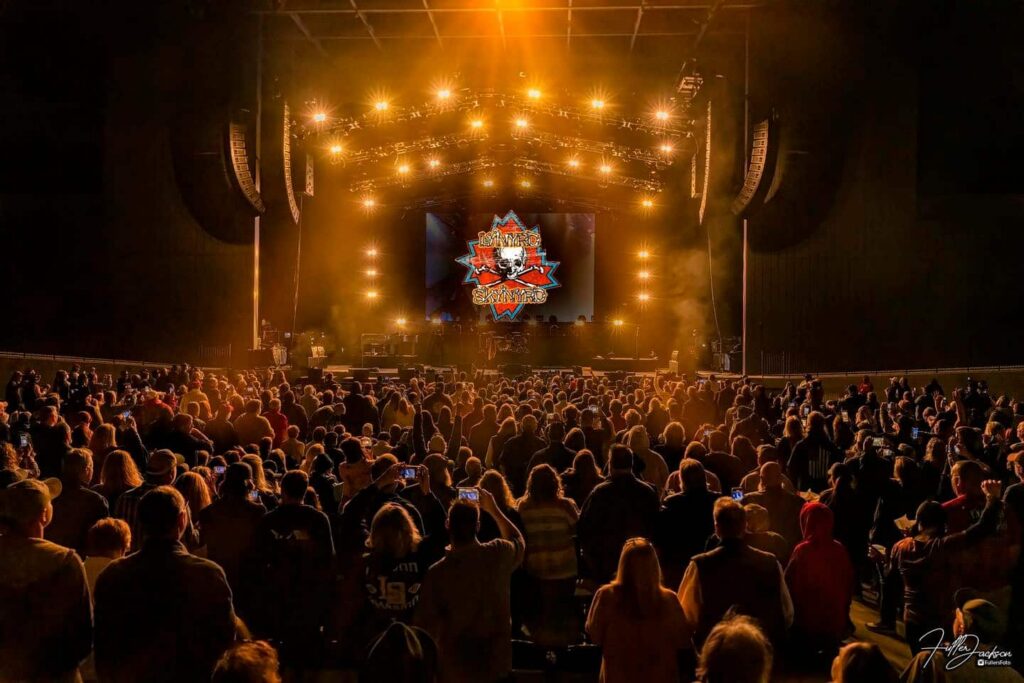 A large audience looks toward a vibrant, illuminated concert stage displaying a graphic logo on the screen. Warm yellow lights fill the venue as numerous spectators capture photos or record videos.