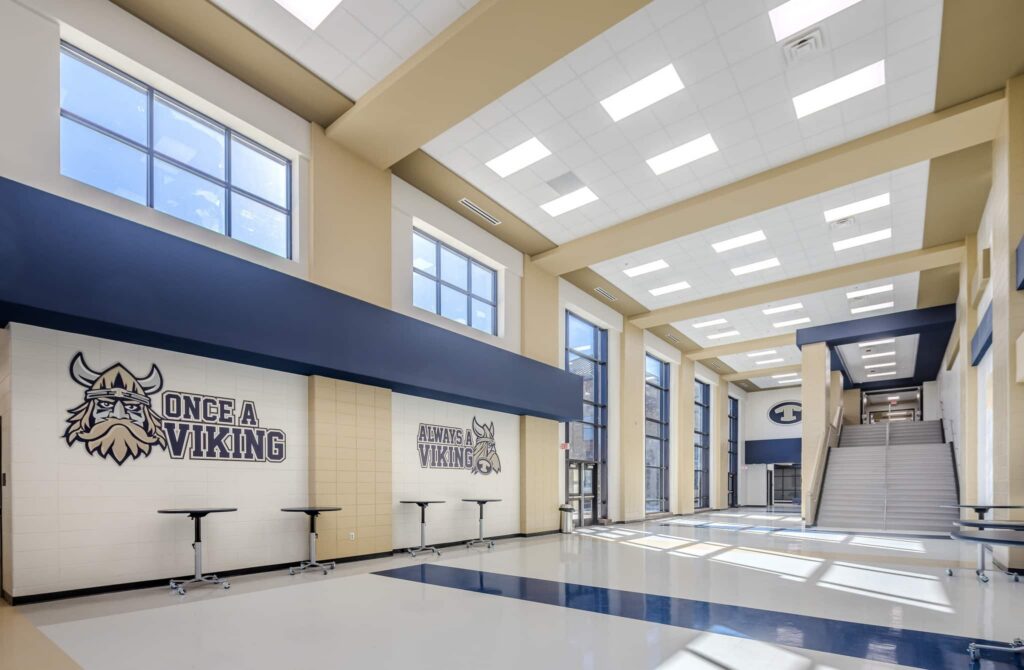A bright school hallway features large windows, blue and beige walls, Viking-themed murals, several tall tables, and a staircase that leads to the upper floor. Sunlight streams across the shiny floor.