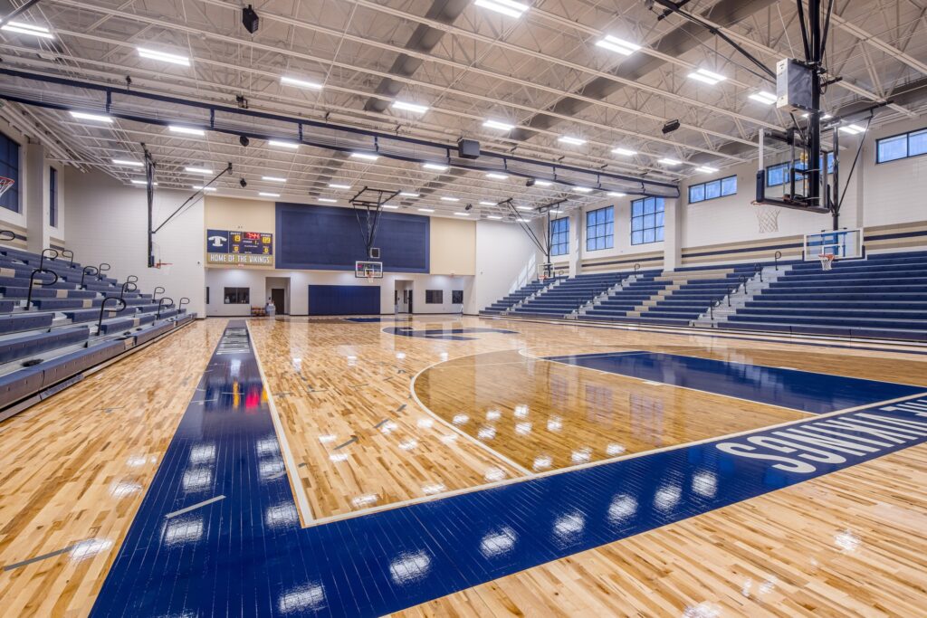 A modern indoor basketball court featuring polished wooden floors, blue and white bleachers lining both sides, basketball hoops at each end, and a scoreboard mounted on the far wall. The well-lit gymnasium is currently empty.