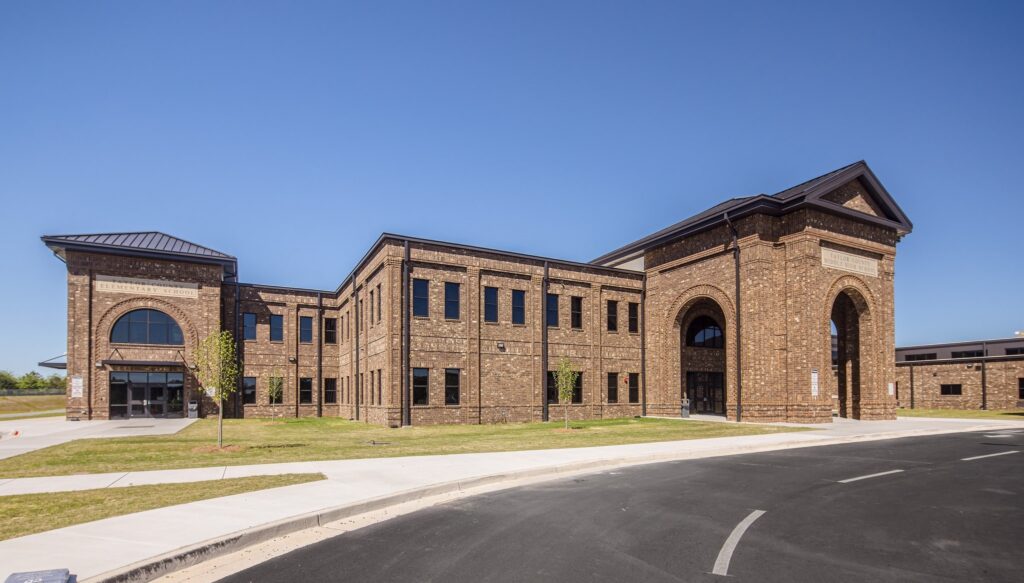 A spacious, contemporary two-story brick building featuring tall arched entrances and numerous windows, set beneath a clear blue sky, with a paved road and grassy area in the foreground.