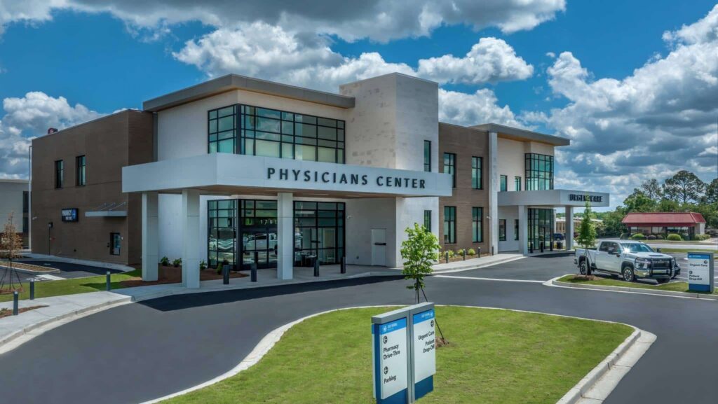 Modern two-story building labeled Physicians Center with large windows, a covered entrance, parked vehicles, and a directional sign in front, surrounded by grass and a partly cloudy sky.