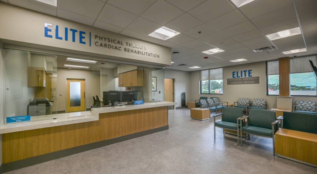 Reception area of a physical therapy and cardiac rehab clinic with chairs, a front desk, signage reading Elite, and large windows letting in natural light. The space is clean, modern, and well-lit.