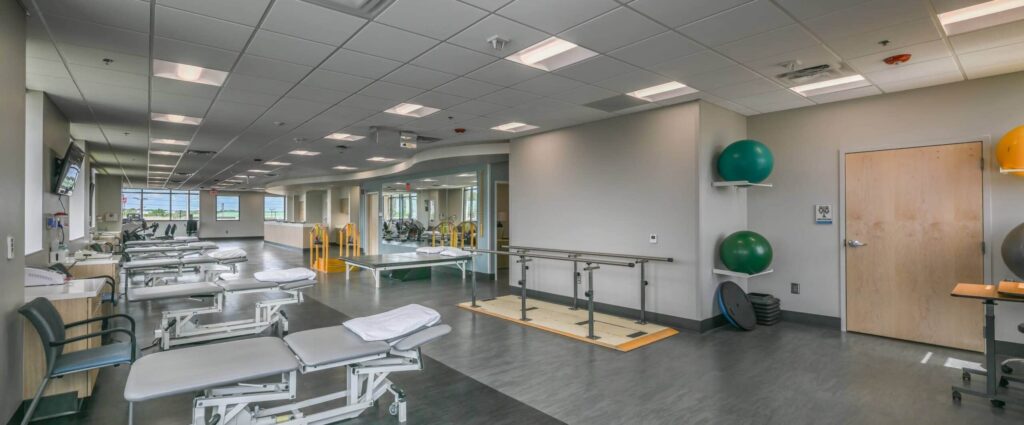 A modern physical therapy clinic featuring treatment tables, exercise machines, parallel bars, therapy balls, and wall mirrors. The space is filled with natural light from large windows and has a tidy, organized look.