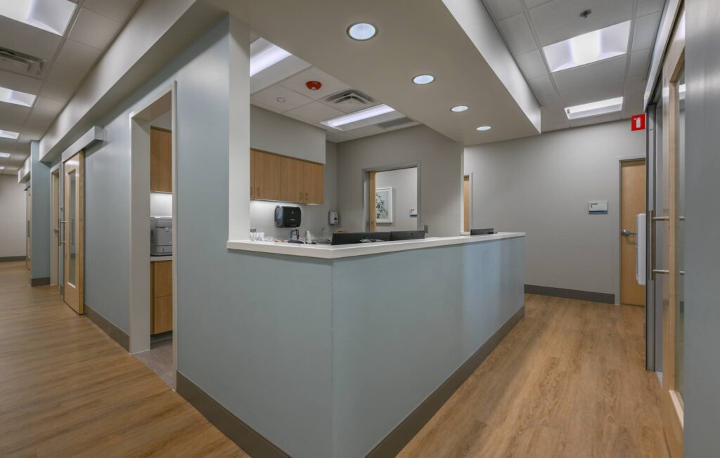 A clean, modern medical office features a central reception desk, light wood flooring, beige walls, and fluorescent ceiling lights. Multiple doors lead to exam rooms.