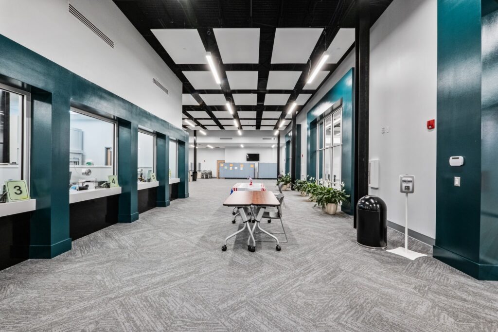A modern office lobby featuring teal walls, several numbered service windows, a central table surrounded by chairs, green plants placed throughout, and a TV mounted on the back wall. The space appears open, well-lit, and tidy.