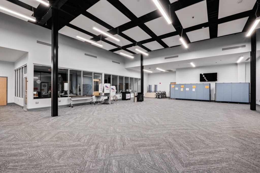 A spacious, modern room featuring gray carpet, glass walls along one side, blue partitions, a TV, tables and chairs arranged throughout, and ceiling lights. The setting suggests it is a multi-purpose or conference room.