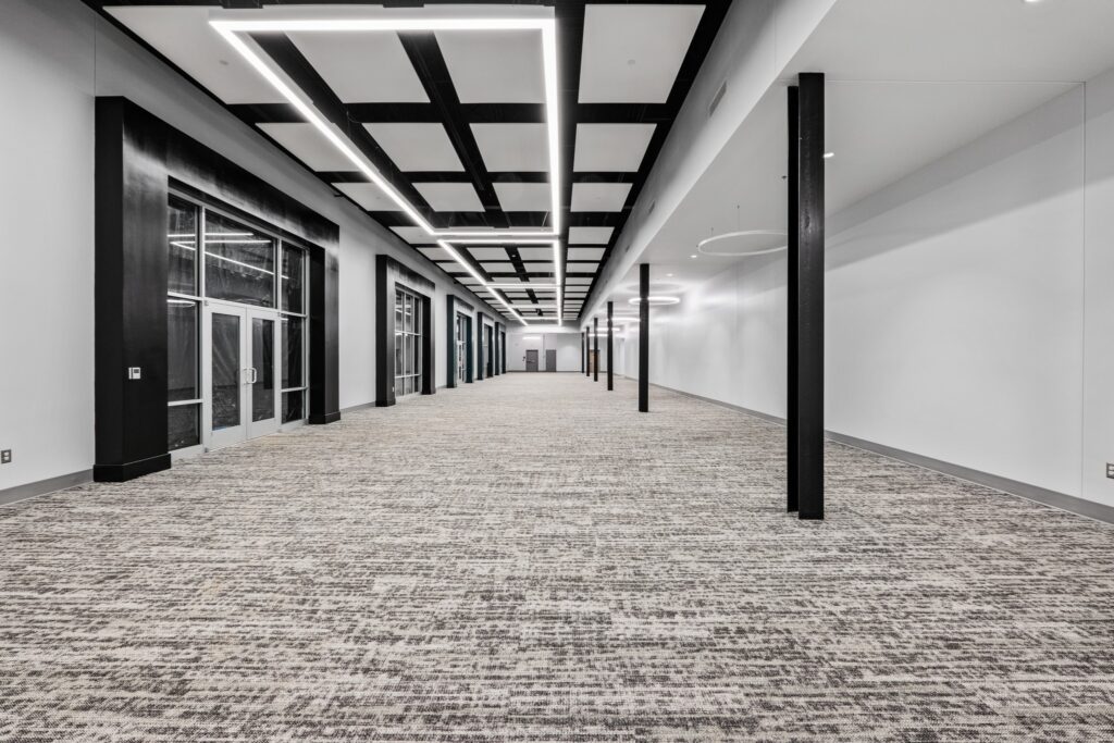 Wide, modern hallway with patterned carpet, black ceiling beams, geometric lighting, floor-to-ceiling windows on the left, and a white wall on the right. The corridor appears empty and well-lit.