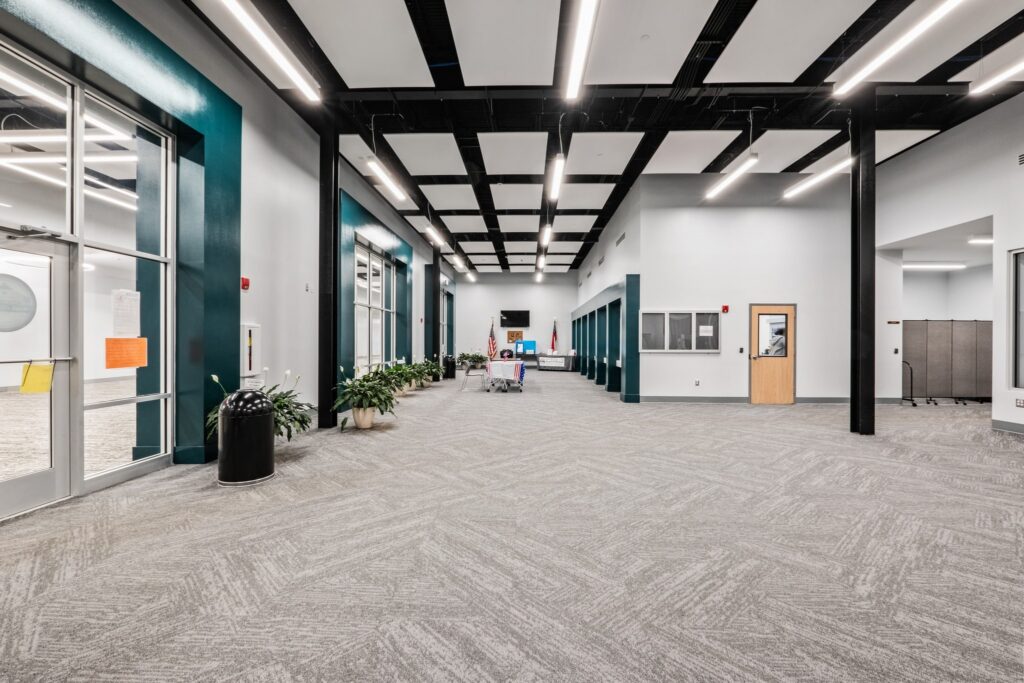 A spacious, modern hallway featuring gray carpet, teal accents, and plants arranged along the walls under bright ceiling lights. Multiple doors and windows line both sides of the hall, with a flag visible at the far end.
