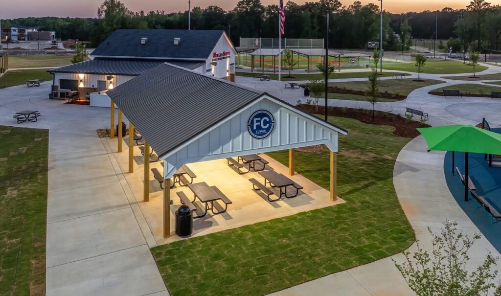 A covered pavilion with picnic tables glows with lights at dusk in a park. Close by are a playground, a baseball field, winding walking paths, and an adjacent building. The entire scene is surrounded by trees and grass.