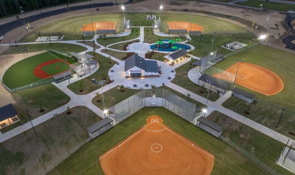 Aerial view showcasing a brightly illuminated baseball and softball complex, multiple fields arranged around a central building, interconnected walkways, and lush green grass covering the entire area.