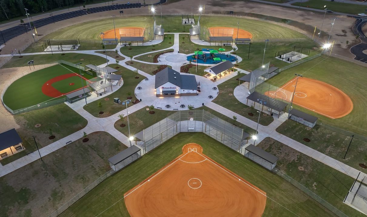 Aerial view showcasing a brightly illuminated baseball and softball complex, multiple fields arranged around a central building, interconnected walkways, and lush green grass covering the entire area.