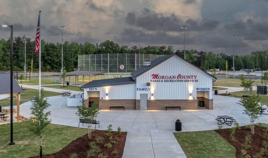 Restroom building at Morgan County Parks & Recreation Services, with Men, Women, and Family entrances. Picnic tables, flagpole, and baseball field are visible in the background under a cloudy sky.