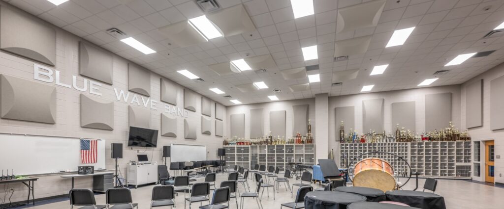 A spacious band room with chairs, music stands, percussion instruments, storage cubbies, trophies, a whiteboard, an American flag, and BLUE WAVE BAND on the wall.