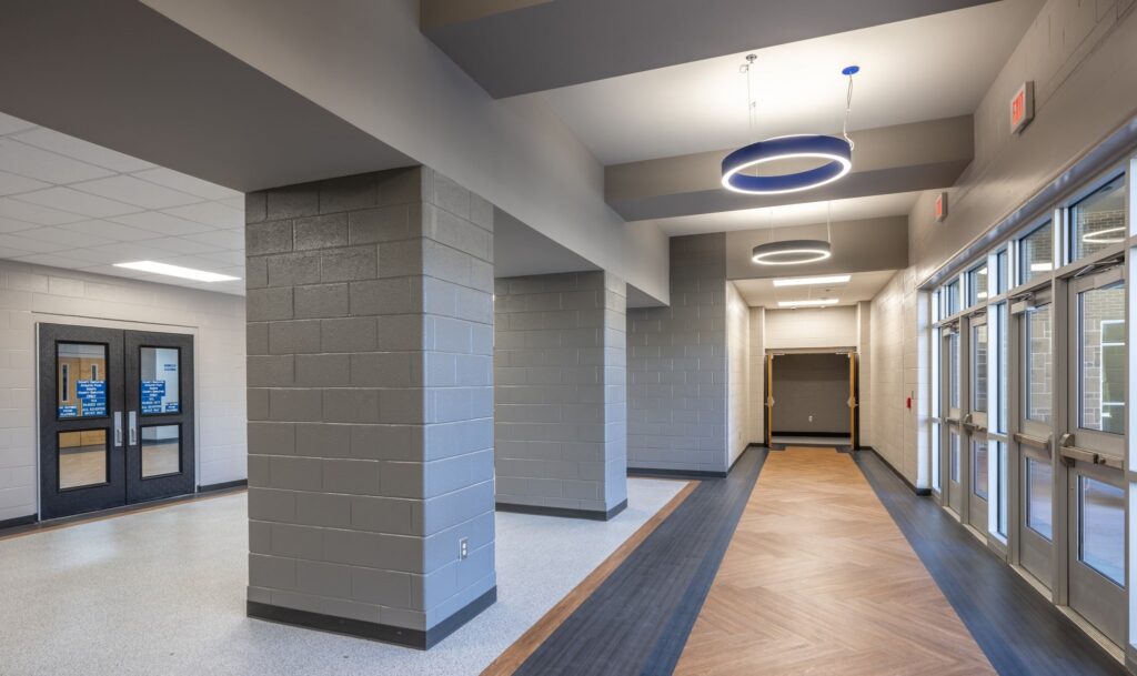 A modern school hallway with gray walls, large pillars, glass doors, wood-patterned flooring, and circular ceiling lights. Natural light enters through the windows along one side.