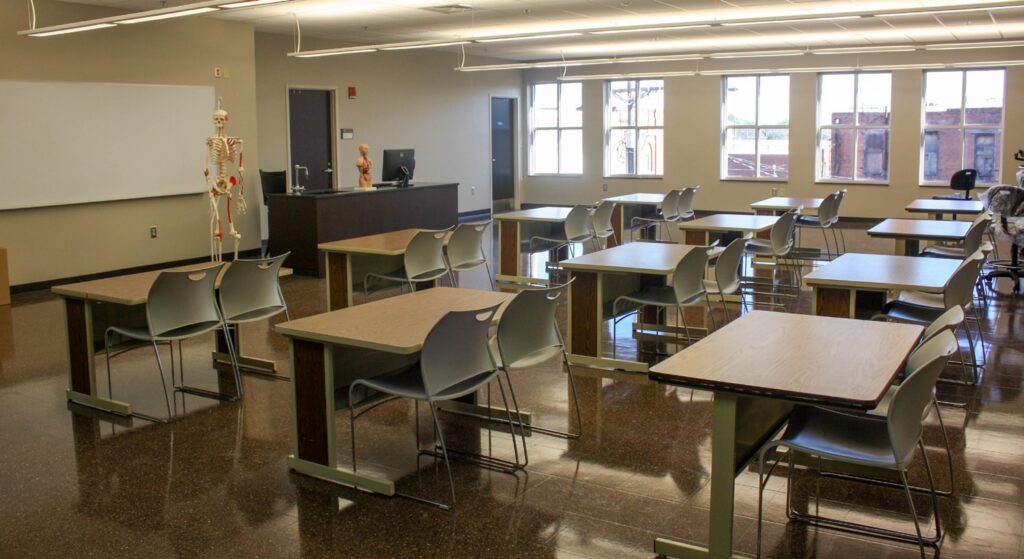 A classroom with empty desks, chairs, and a teacher’s desk. A human skeleton model stands near the board, and anatomical models are on the teacher’s desk. Large windows let in natural light.