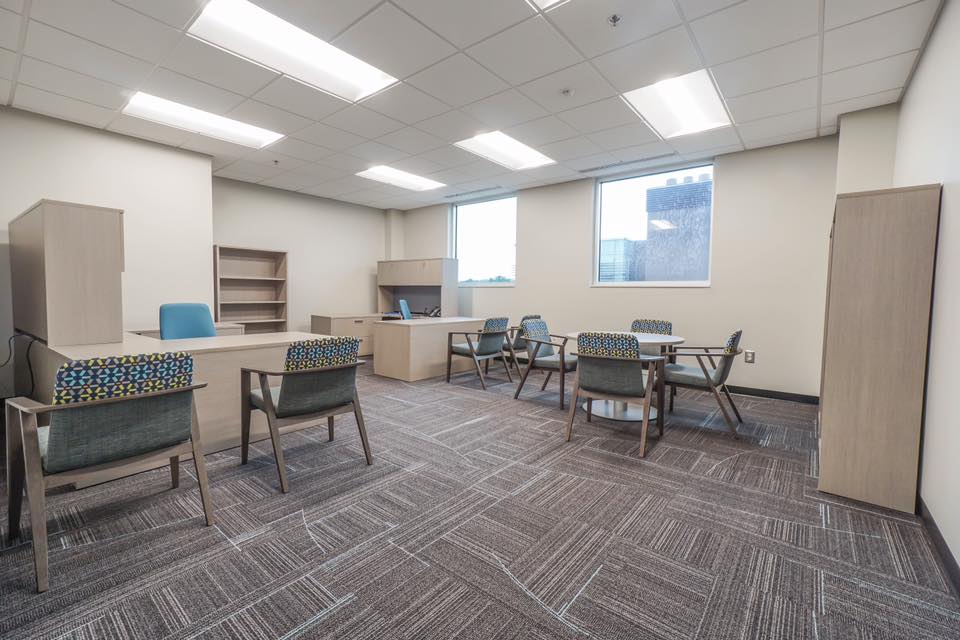 A modern office with desks, chairs, shelves, and large windows letting in natural light. The space features neutral colors, open seating, and carpeted flooring.