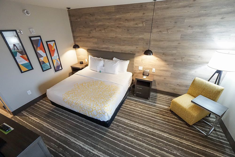 A modern hotel room featuring a double bed with a yellow-patterned bedspread, two nightstands, pendant lights, a yellow armchair, floor lamp, striped carpet, and abstract art on the wall.