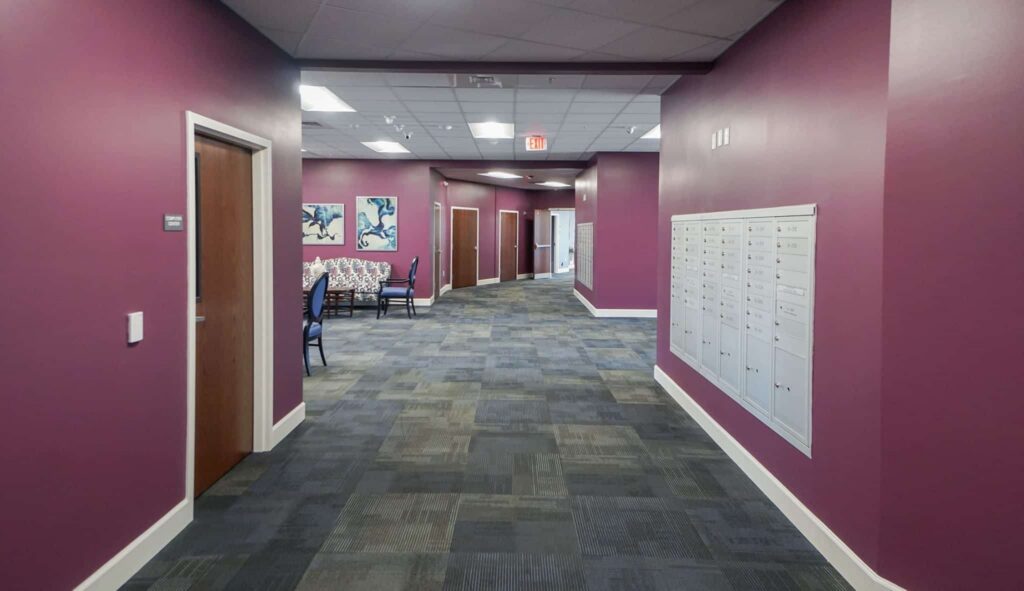 A hallway featuring purple walls and gray carpet, lined with a row of mailboxes and framed artwork. Closed wooden doors are visible, and at the far end, there is a seating area furnished with patterned chairs.