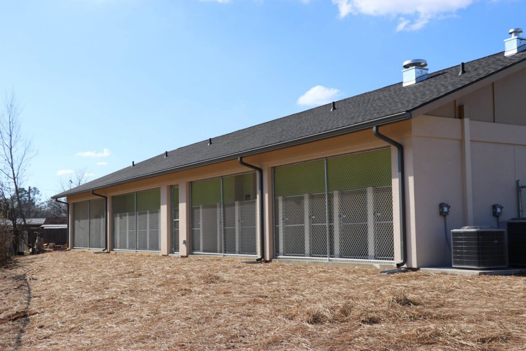 A structure with a slanted roof and multiple spacious fenced areas, possibly designed for animals, stands on straw-covered ground beneath a blue sky dotted with scattered clouds.