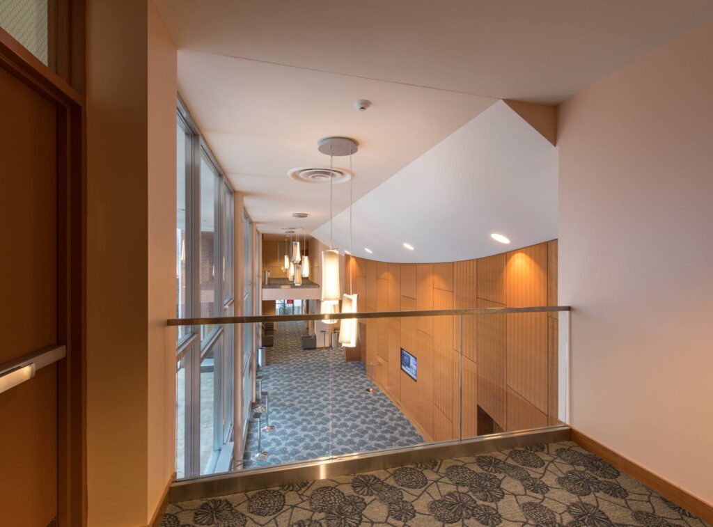 A modern hallway with patterned carpet, wooden walls, hanging pendant lights, large windows lining the left side, and a glass railing providing a view of the lower level.