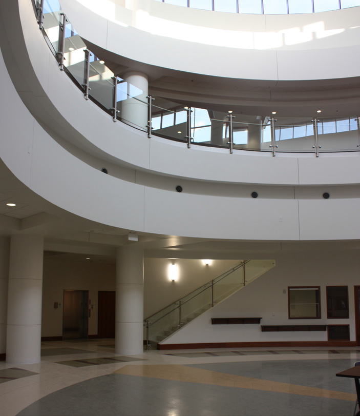 A spacious modern building interior features curved white balconies, glass railings, and a staircase. Sunlight pours in through large windows above, illuminating the open, multi-story atrium.