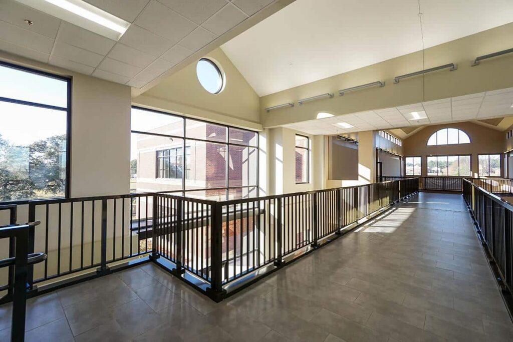 Bright, modern indoor walkway featuring large windows, metal railings, and tiled flooring. Sunlight pours through the glass, lighting up the open area and offering views of surrounding trees and neighboring buildings.